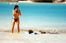 A shy thin pale white chick with jet black hair stands topless on a beach. She has a..